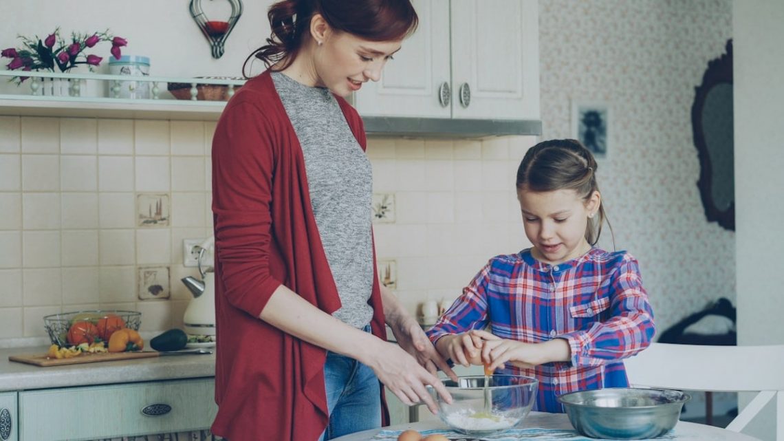 Little cheerful girl helping her mother in the kitchen cropping egg into bowl and mixing dough for cookies. Family, food, home and people concept