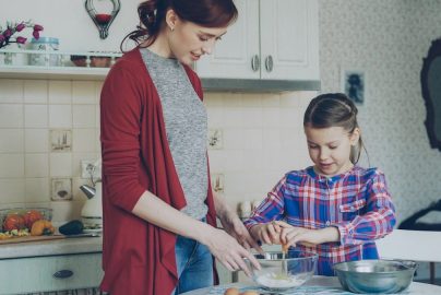 Little cheerful girl helping her mother in the kitchen cropping egg into bowl and mixing dough for cookies. Family, food, home and people concept