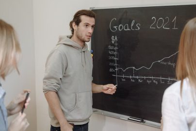 Three professionals engaged in a business discussion about goals written on a chalkboard in an office.